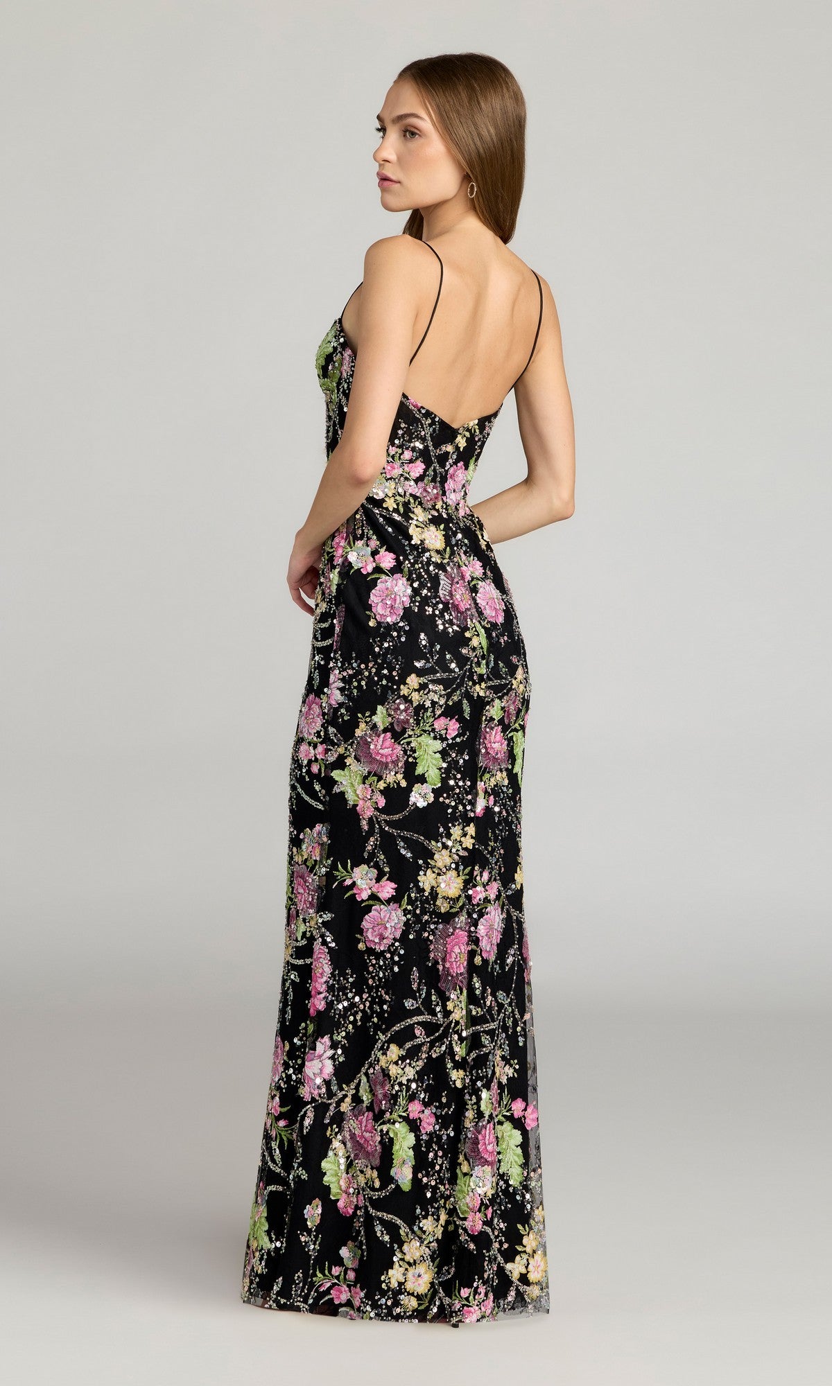 A woman wears the Mac Duggal 68891 Beaded Long Floral Formal Dress, featuring a black floor-length design with spaghetti straps, standing and looking over her shoulder against a neutral background.